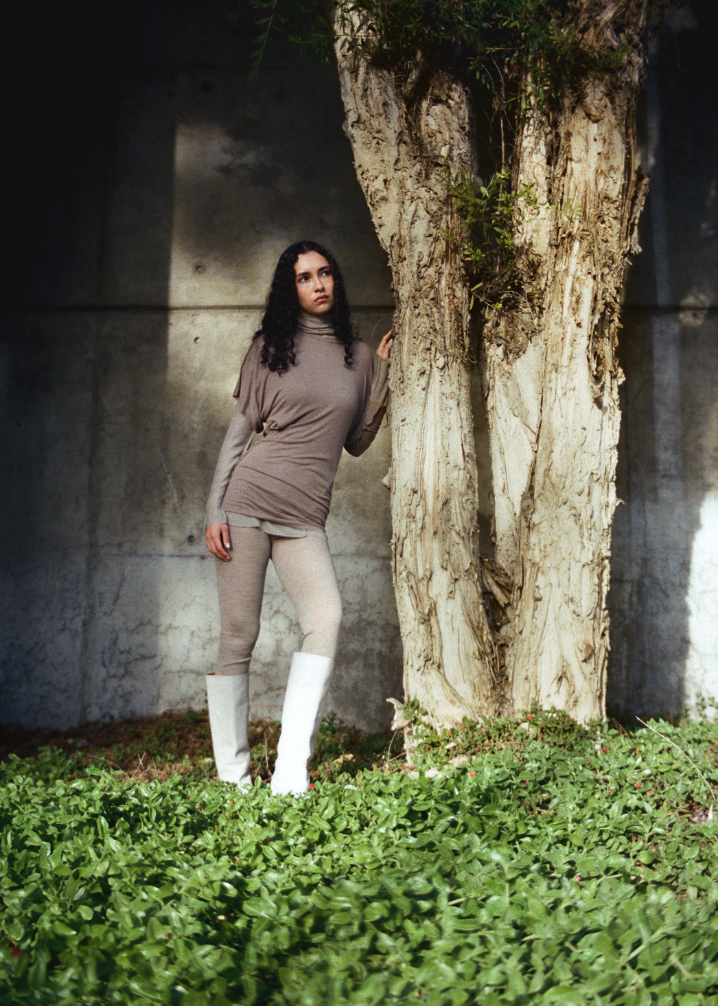 Woman standing next to a tree in a grassy area with a concrete wall in the background