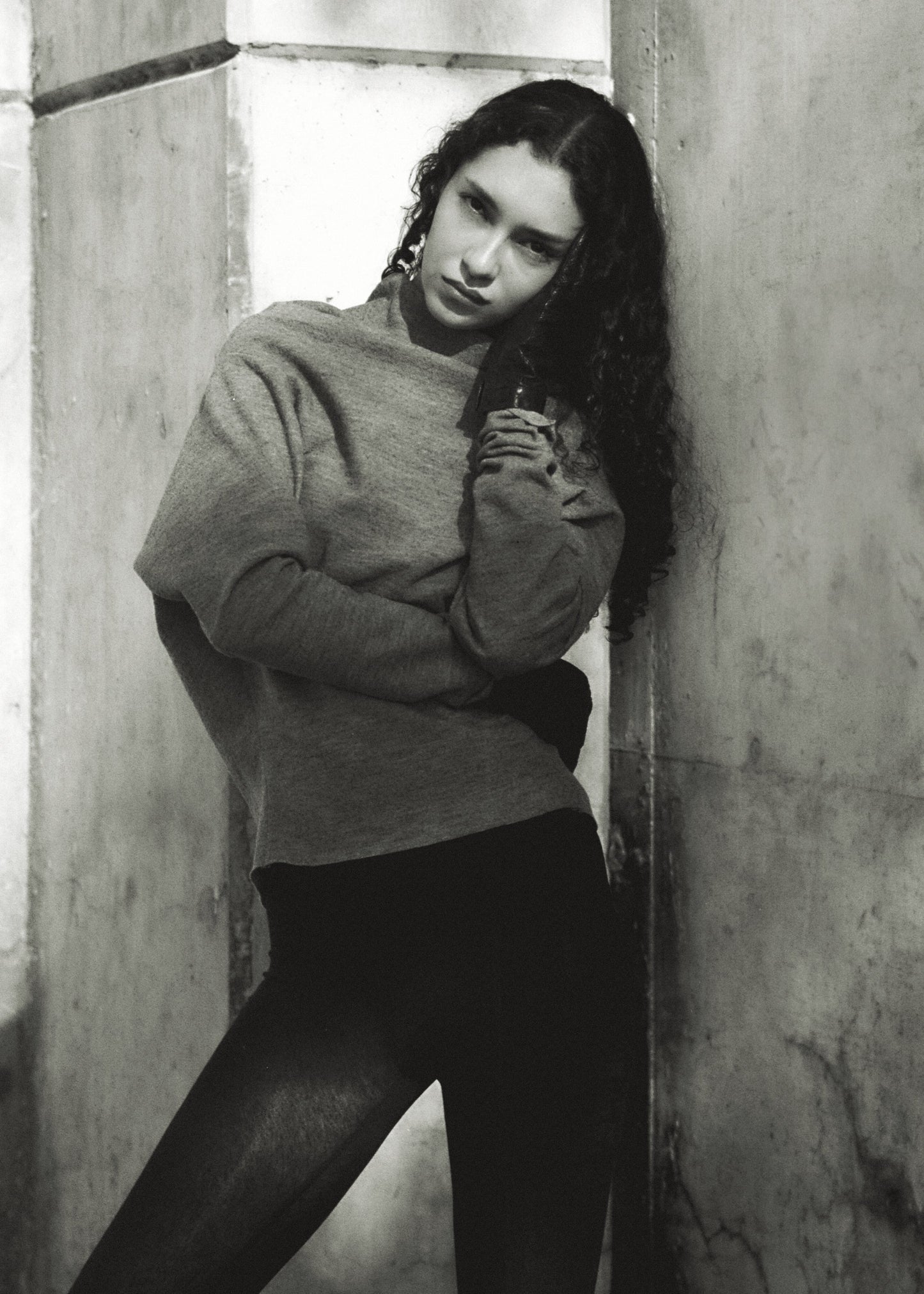 Person wearing a gray sweater and black pants against a concrete wall.
