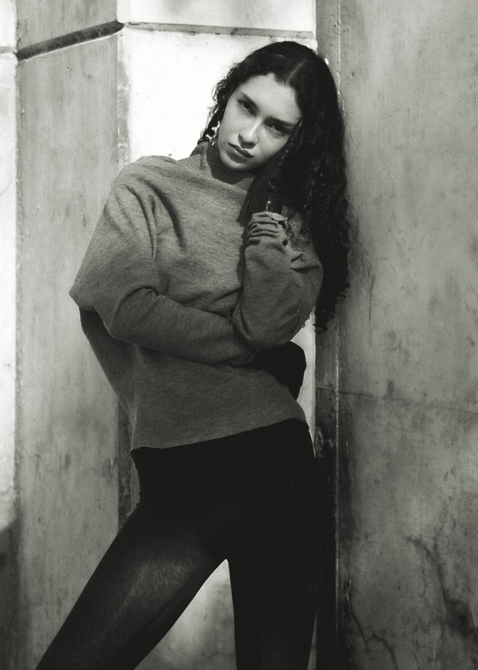 Person wearing a gray sweater and black pants against a concrete wall.